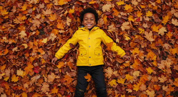 Overhead shot of a black kid smiling in yellow rain jacket and lying in a pile of fall leaves #3