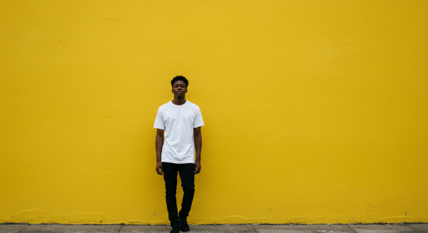 A young black man standing by a yellow wall in a far-off shot #4