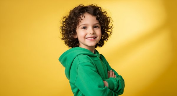 Smiling child with curly hair wearing a green hoodie, standing confidently with arms crossed, bright yellow background #1