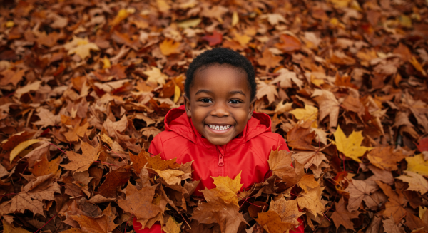 Overhead shot of a black kid smiling in red rain jacket and lying in a pile of fall leaves #3