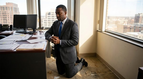 A black man in his professional suit on his knees praying in his office